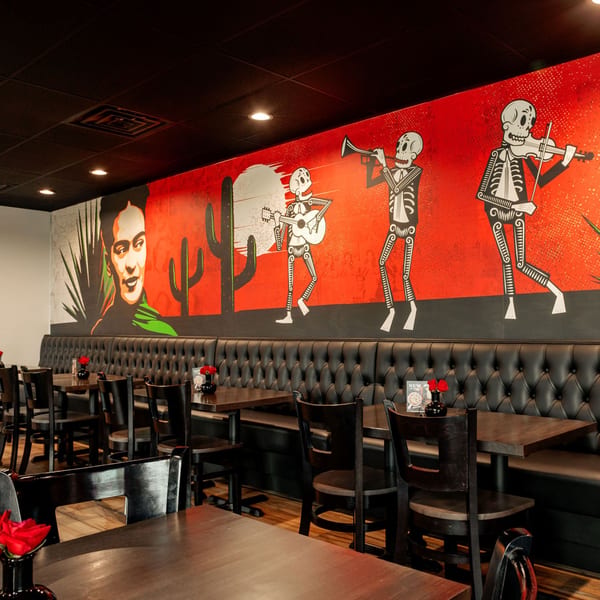 Restaurant interior with Frida Kahlo mural, skeleton musicians, black booths, and wooden tables with red flowers.