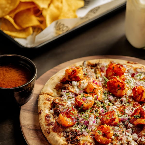 Birria Love's shrimp pizza with red onions and cilantro, served with a side of dipping sauce and tortilla chips.