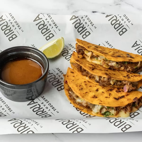 Three Birria tacos with melted cheese and meat, a side of consommé, and a lime wedge.