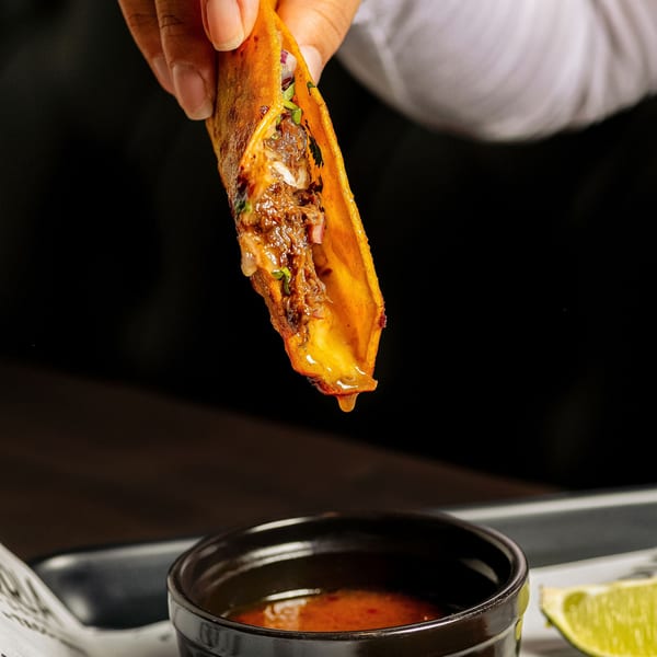 Hand holding a Birria Love taco, dripping consommé into a small black bowl with lime.