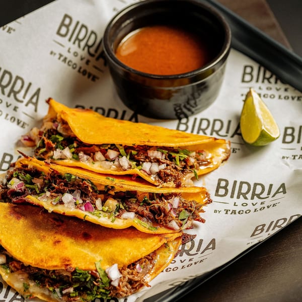 Three Birria Love tacos with shredded meat, onions, cilantro, and melted cheese, with a side of consommé and lime.