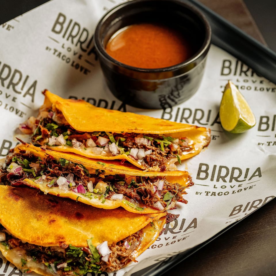 Three Birria Love tacos with shredded meat, onions, cilantro, and melted cheese, with a side of consommé and lime.