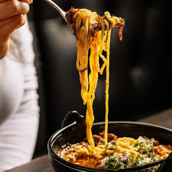 A fork lifts cheesy birria ramen noodles from a black bowl, garnished with cilantro and onions.