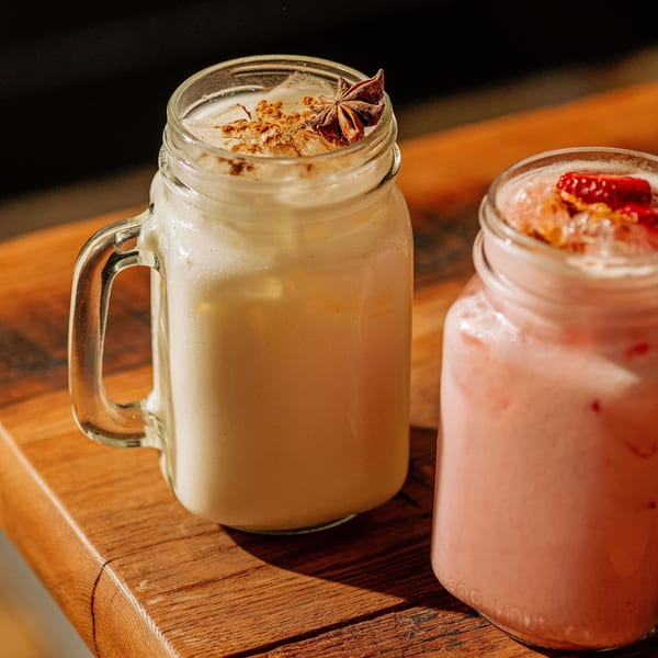Two mason jar drinks on a wooden surface, one horchata with star anise, one pink with fruit.