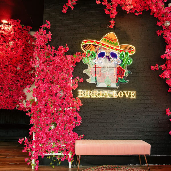 A neon Birria Love sign with a sugar skull and sombrero, surrounded by vibrant pink floral decor.