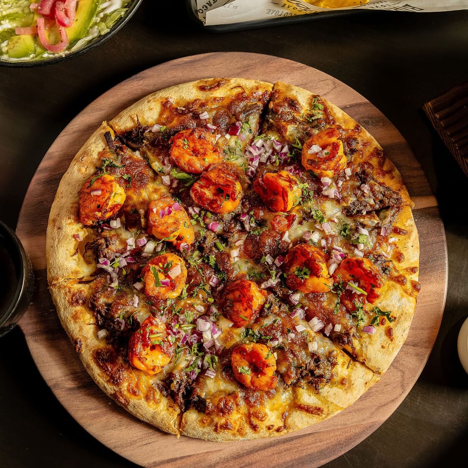 Birria Love's Mexican pizza, topped with shrimp, birria, red onions, and cilantro on a wooden board.