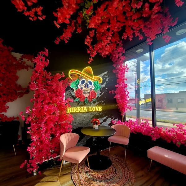 Birria Love restaurant interior with pink floral decor, a neon skull sign, and a small table with chairs.
