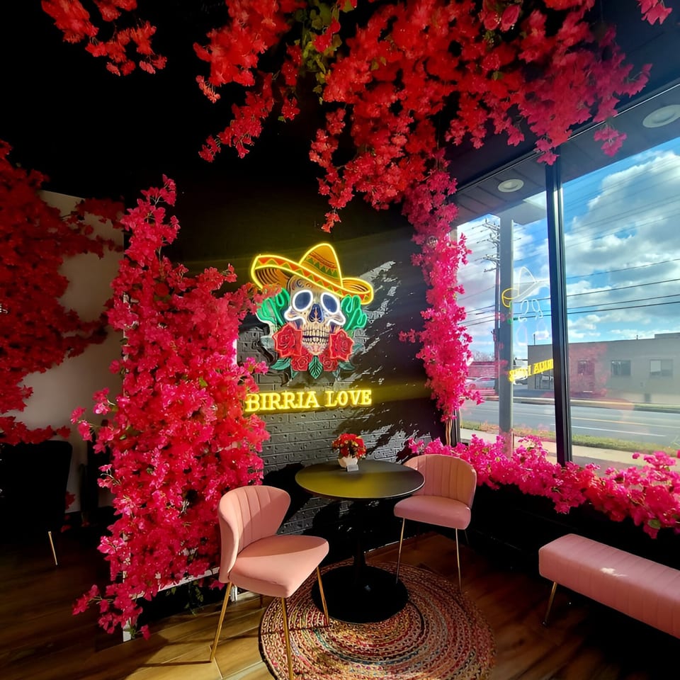 Birria Love restaurant interior with pink floral decor, a neon skull sign, and a small table with chairs.
