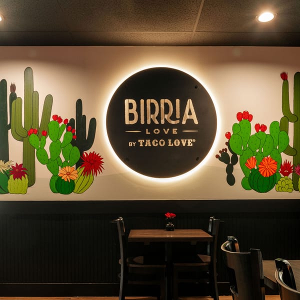 Birria Love restaurant interior with a backlit logo and a colorful cactus mural above dark wood paneling.