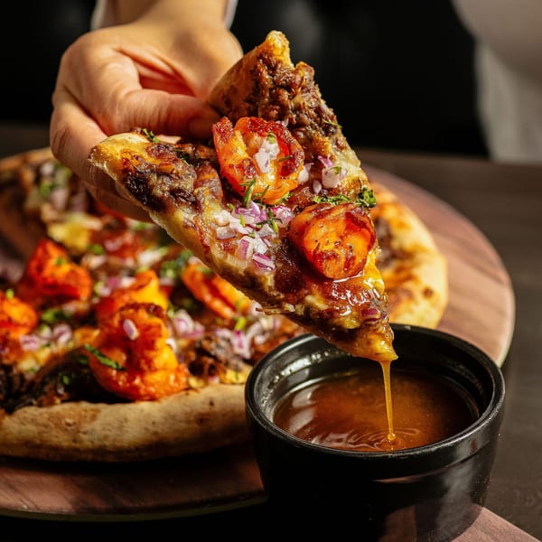 Hand holding a slice of Birria Love pizza with shrimp, dipping it into a small black bowl of sauce.