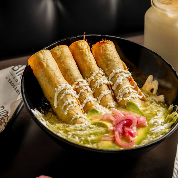 Four golden taquitos drizzled with white sauce and cheese, served in a black bowl with green salsa, avocado, and pickled onions.