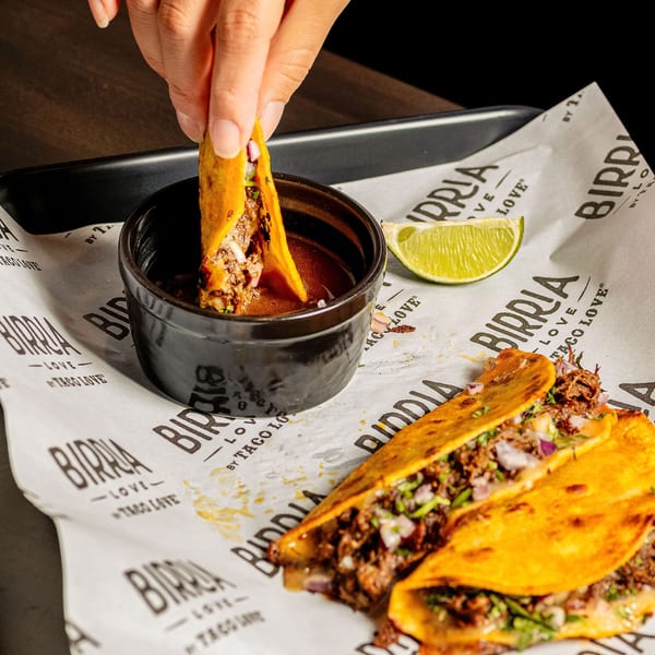 A hand dips a Birria Love taco into a small black bowl of consommé, with two other tacos and a lime wedge on branded paper.